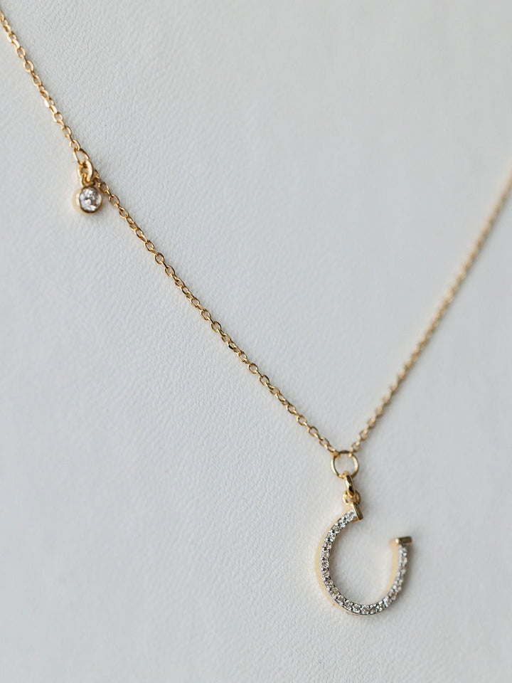 The Savannah Sevens western life{&}style Diamond Horseshoe Necklace features a delicate gold chain with a small round diamond charm and a 14K gold horseshoe pendant encrusted with sparkling stones, shown on a light background.