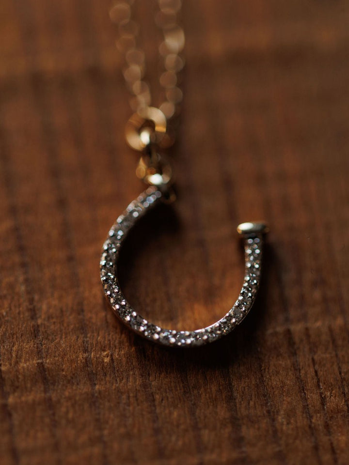 A close-up of the Savannah Sevens western life{&}style Diamond Horseshoe Necklace, with a 14K gold chain and diamond-encrusted horseshoe pendant, shown resting on a wooden surface.