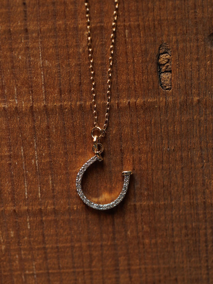 The Diamond Horseshoe Necklace by Savannah Sevens western life{&}style features a sparkling horseshoe pendant, shown on a wooden surface.