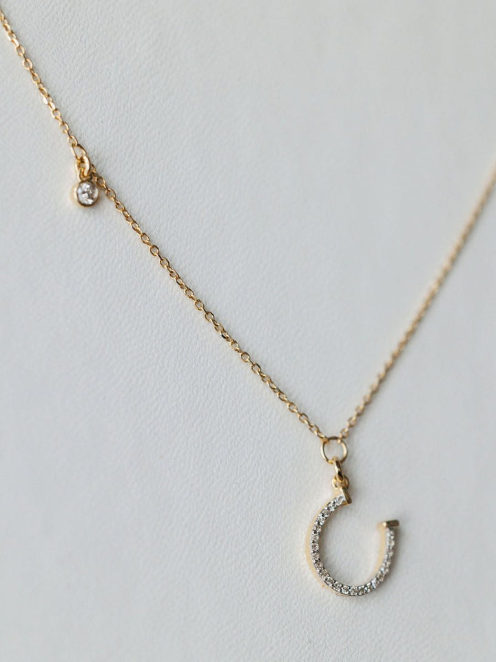 The Savannah Sevens western life{&}style Diamond Horseshoe Necklace features a delicate gold chain with a small round diamond charm and a 14K gold horseshoe pendant encrusted with sparkling stones, shown on a light background.