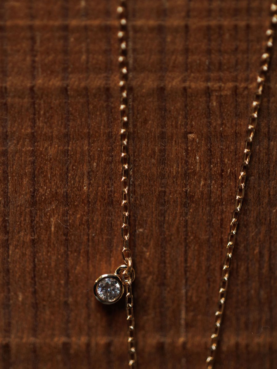 A close-up of the Diamond Horseshoe Necklace by Savannah Sevens western life{&}style, featuring a small, round diamond pendant on a 14K gold chain, displayed on a textured wooden surface.