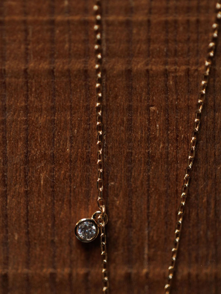 A close-up of the Diamond Horseshoe Necklace by Savannah Sevens western life{&}style, featuring a small, round diamond pendant on a 14K gold chain, displayed on a textured wooden surface.