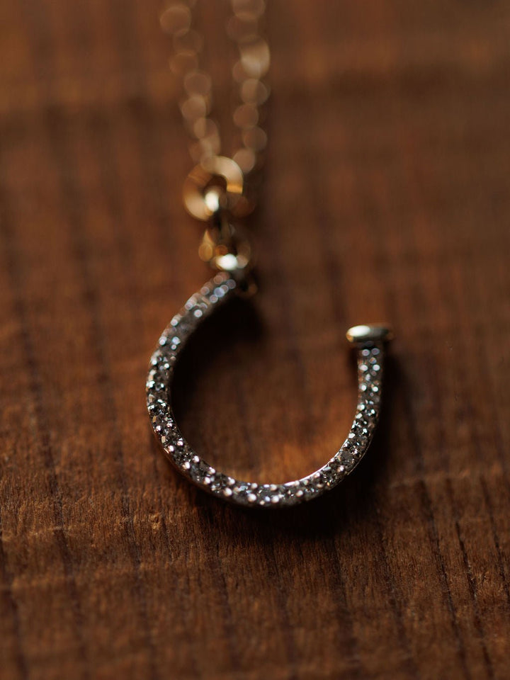 A close-up of the Savannah Sevens western life{&}style Diamond Horseshoe Necklace, with a 14K gold chain and diamond-encrusted horseshoe pendant, shown resting on a wooden surface.
