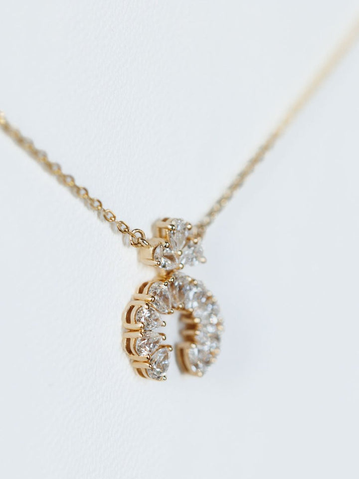 A close-up of the Savannah Sevens western life{&}style Diamond Squash Necklace in 14K gold, featuring a crescent pendant adorned with round and pear-shaped diamonds, displayed on a white background.