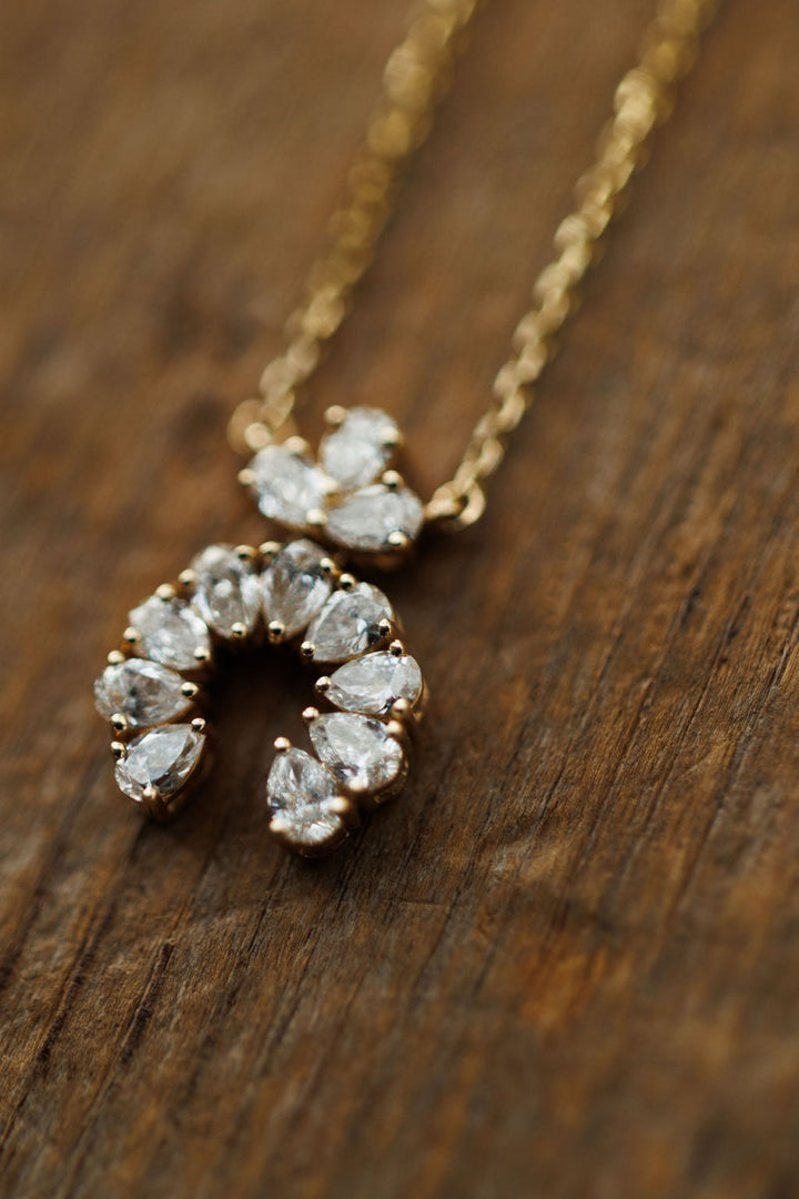 The Diamond Squash Necklace by Savannah Sevens western life{&}style features a 14K gold pendant with sparkling pear-shaped diamonds, shaped like a horseshoe and displayed on a wooden surface.