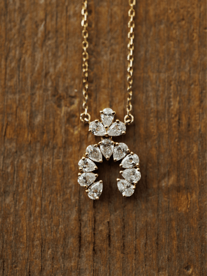 The Diamond Squash Necklace by Savannah Sevens western life{&}style features clear, pear-shaped diamonds in a horseshoe and fan pattern pendant, crafted in 14K gold and displayed on a wooden surface.