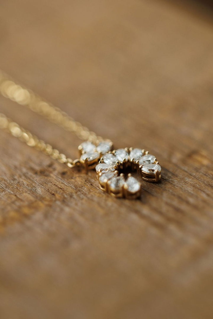 The Diamond Squash Necklace by Savannah Sevens western life{&}style features a 14K gold horseshoe-shaped pendant adorned with sparkling diamonds, elegantly displayed on a wooden surface.