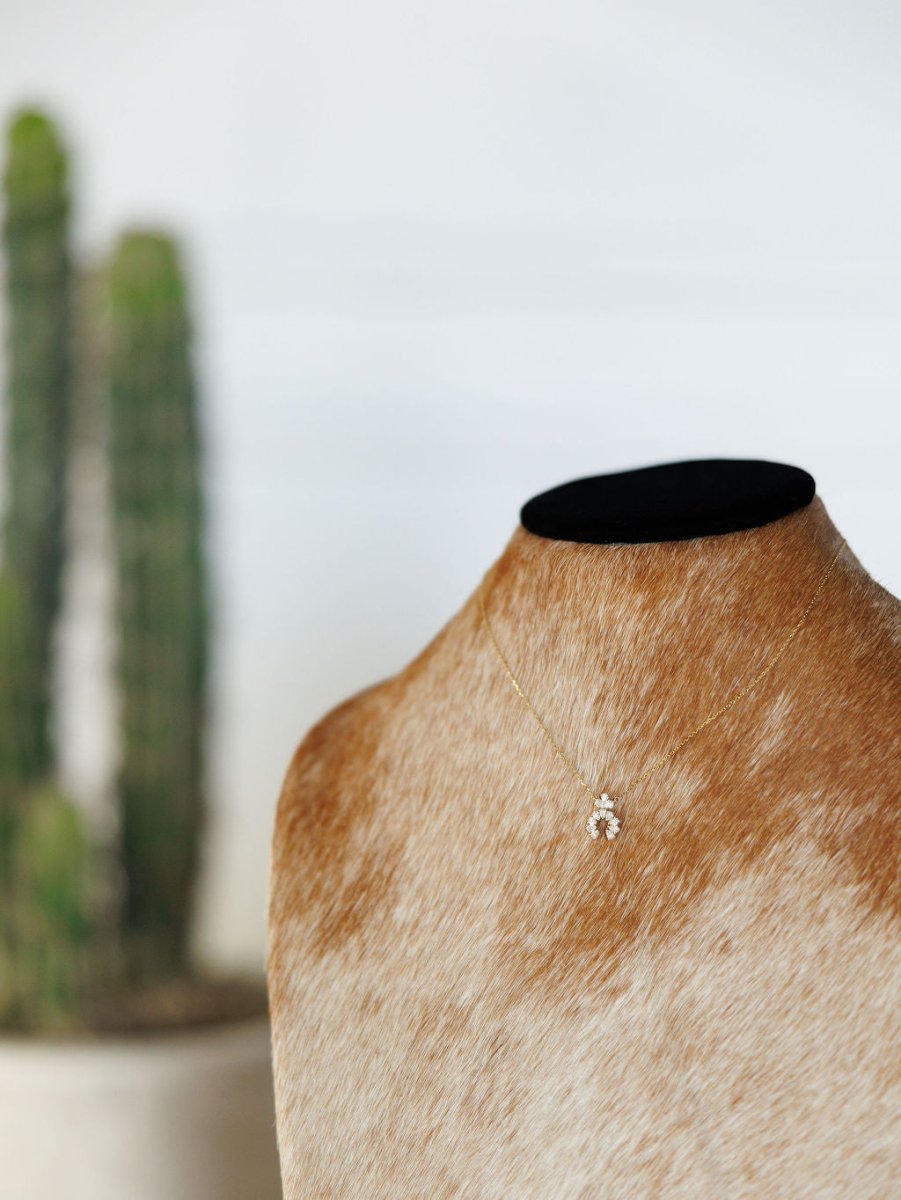The Diamond Squash Necklace by Savannah Sevens western life{&}style is a delicate 14K gold piece with a small pendant, shown on a brown cowhide bust with a blurred green cactus in the background.