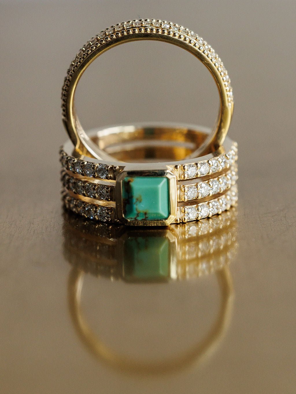 A close-up of Savannah Sevens western life{&}style’s Diamond Turquoise Ring with lab grown diamonds and a rectangular turquoise stone beside a simple beaded-edge band, both displayed on a reflective surface. Collaboration the Devereaux Gold.
