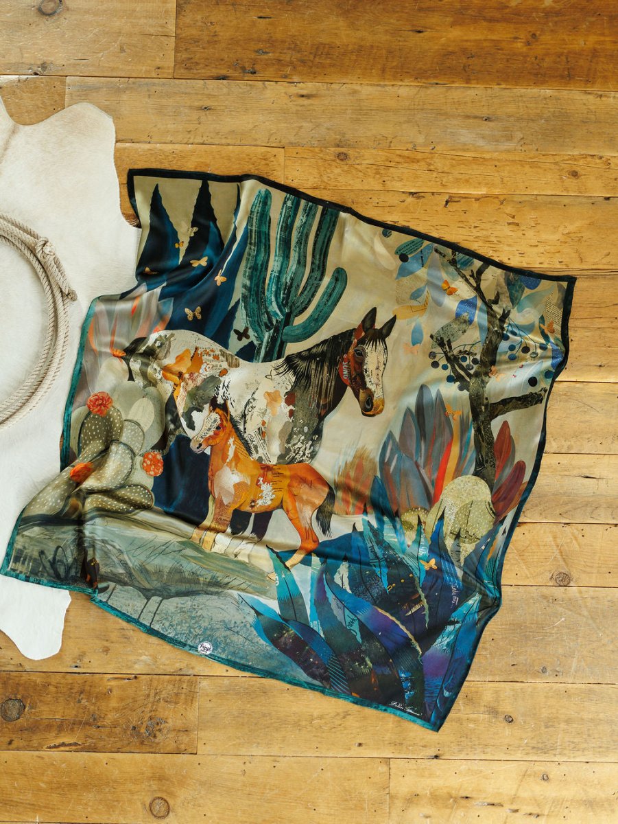 The Dolan Wild Rag by Savannah Sevens western life{&}style features two horses amid cacti, trees, and abstract foliage in a vibrant silk design inspired by Dolan Geiman's Southwest horse scarf. Shown on a wooden floor next to a white cowhide rug.