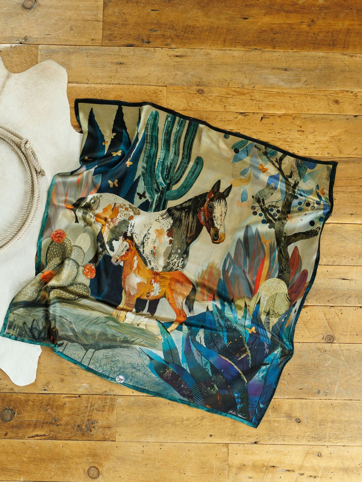 The Dolan Wild Rag by Savannah Sevens western life{&}style features two horses amid cacti, trees, and abstract foliage in a vibrant silk design inspired by Dolan Geiman's Southwest horse scarf. Shown on a wooden floor next to a white cowhide rug.