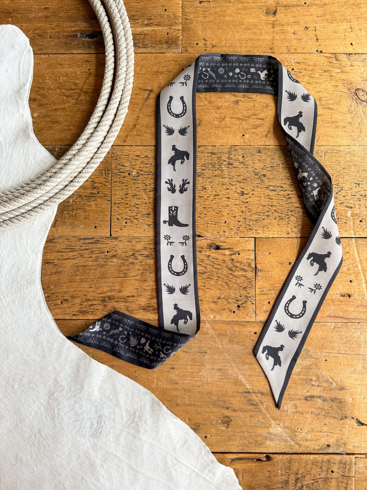 The Down South Black Satin Twilly by Savannah Sevens western life{&}style, adorned with cowboy-inspired motifs, is displayed on a wooden surface beside a white cowhide and a coiled rope.