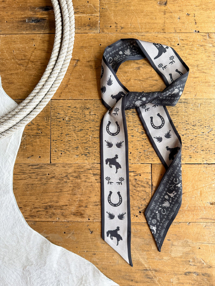 The Down South Black Satin Twilly by Savannah Sevens western life{&}style, featuring black-and-white western prints with horses and horseshoes, is tied on a wooden floor next to a coiled rope and a white cowhide rug.