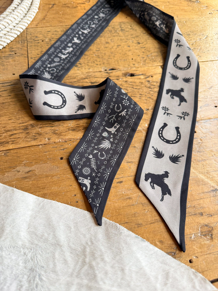 The Down South Black Satin Twilly by Savannah Sevens western life{&}style, featuring black and gray Western-themed prints with horses, horseshoes, and stars, lies on a wooden surface next to a white textured fabric.