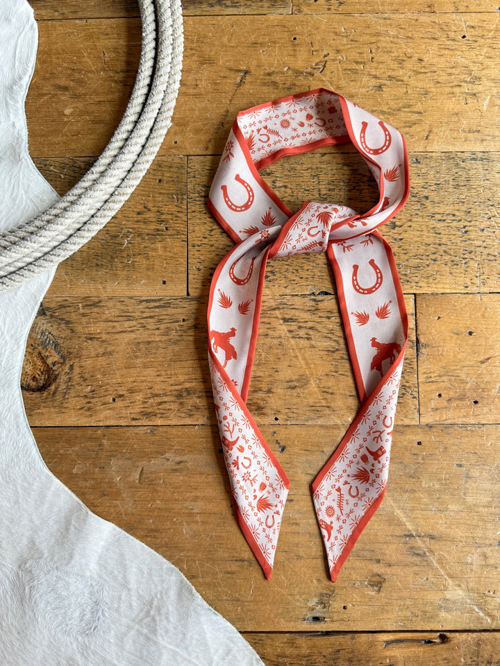 The Down South Burnt Orange Satin Twilly from Savannah Sevens western life{&}style, featuring horseshoe, cactus, and cowboy-inspired prints, lies on a wooden floor next to a coiled rope and a white cowhide rug. Crafted from satin poly chiffon, this twilly adds a stylish Western flair to any outfit.