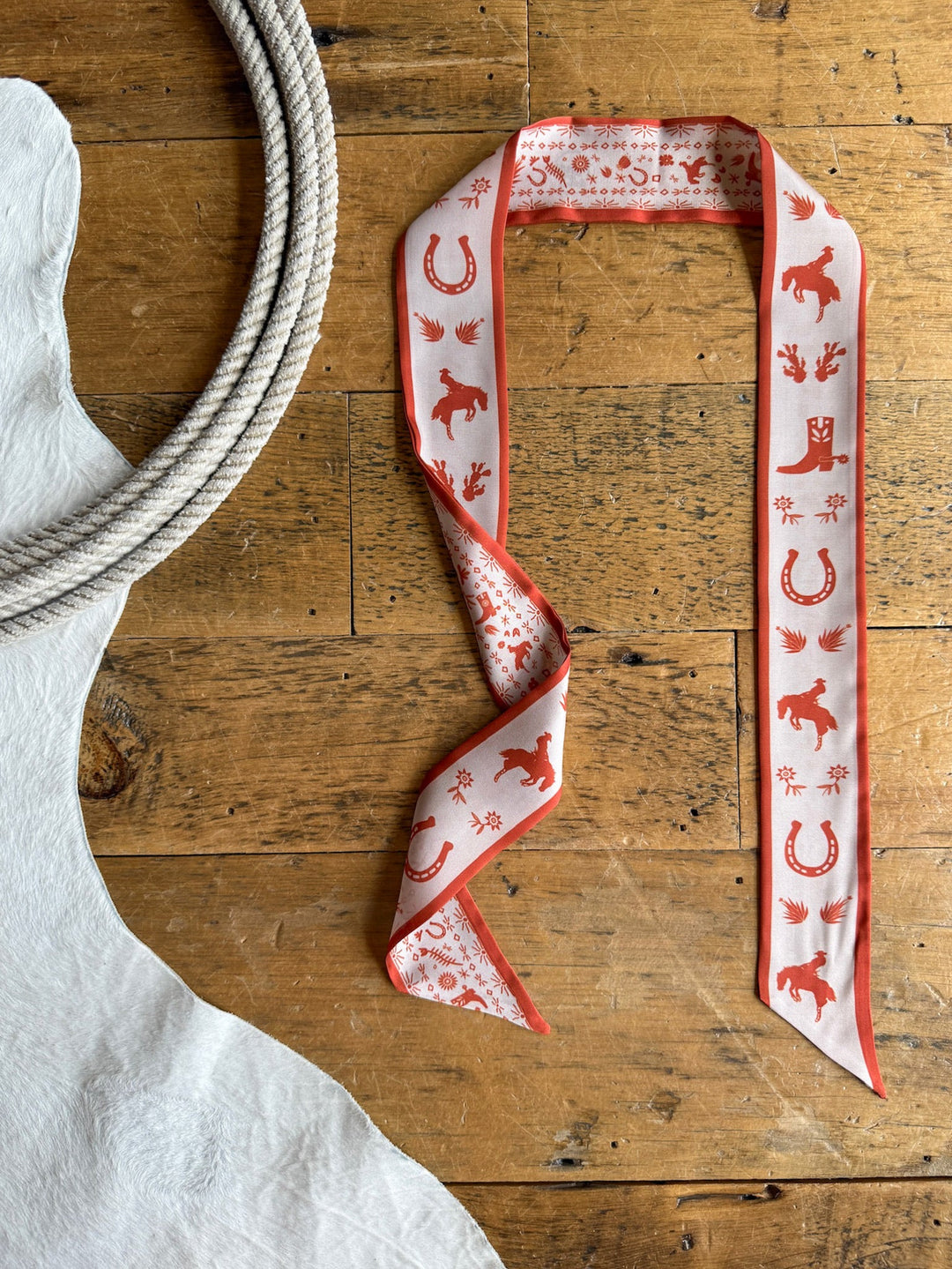 A Down South Burnt Orange Satin Twilly from Savannah Sevens western life{&}style lies on a wooden surface next to a coiled rope and a white cowhide rug.