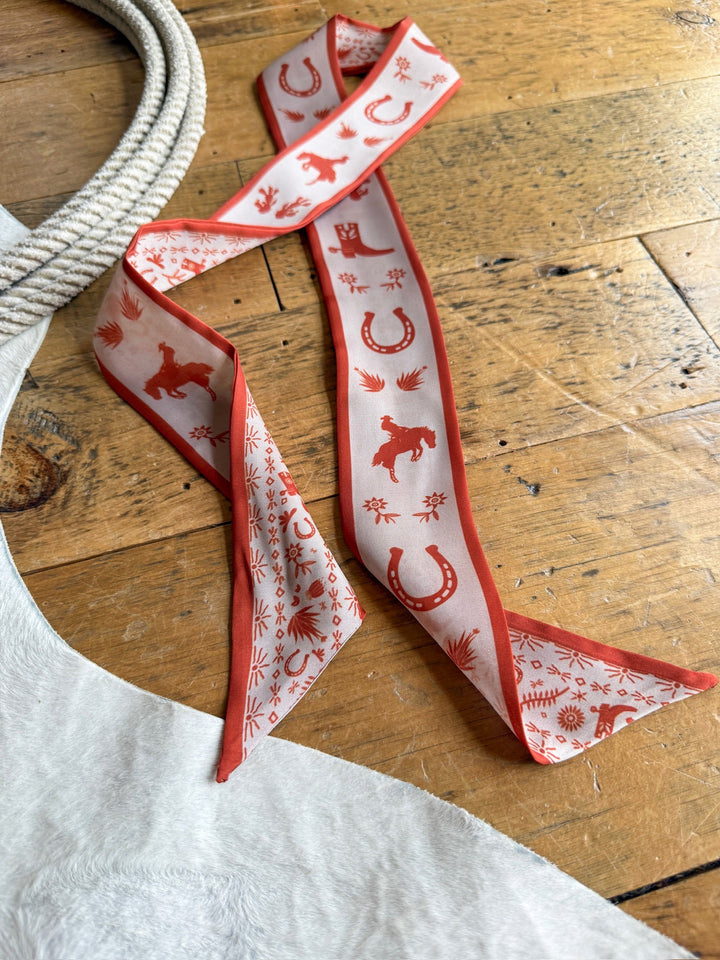 The Savannah Sevens western life{&}style Down South Burnt Orange Satin Twilly, featuring horse, horseshoe, and sun motifs in red and white, is displayed on a wooden floor beside a coiled rope and the edge of a white cowhide rug.