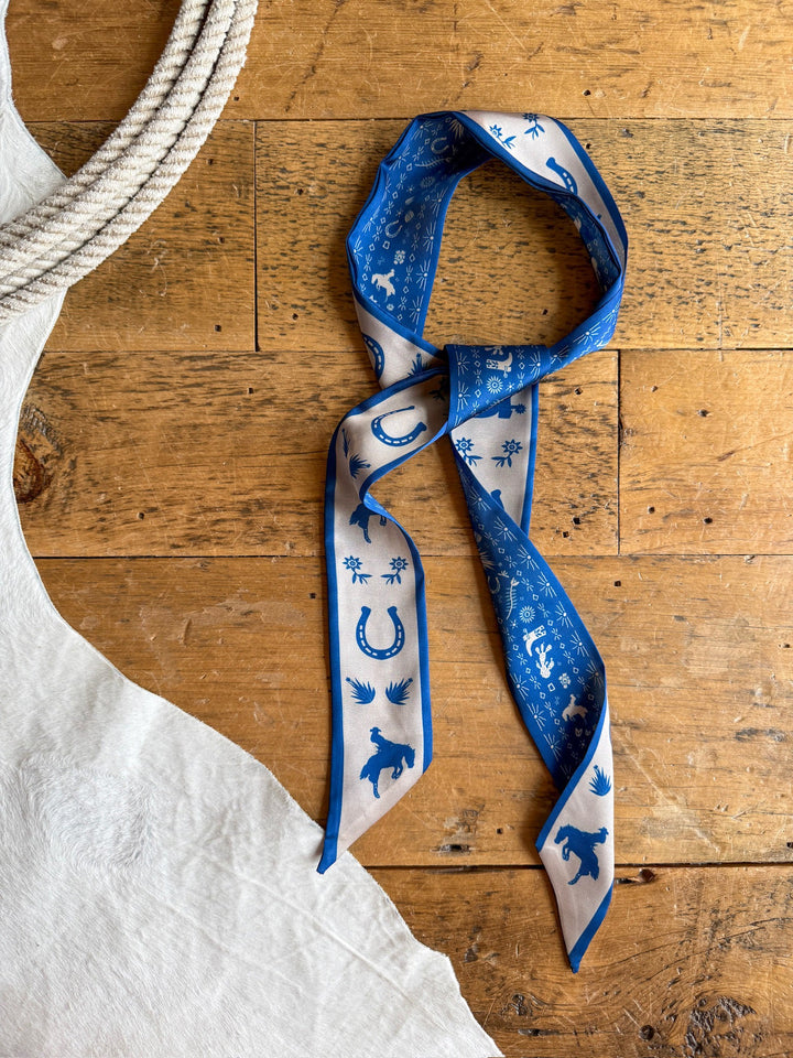 The Down South Cobalt Satin Twilly by Savannah Sevens western life{&}style, featuring blue and white western-themed prints of horses, horseshoes, and plants, is displayed on a wooden surface beside coiled rope and a white textured fabric.