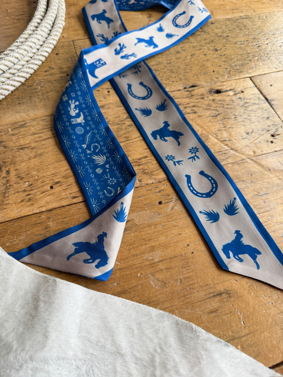 The Down South Cobalt Satin Twilly by Savannah Sevens western life{&}style, featuring a western-inspired print with cowboy, horse, horseshoe, and cactus designs in blue and beige satin, is displayed on a wooden surface beside a coiled rope and a piece of white fabric.