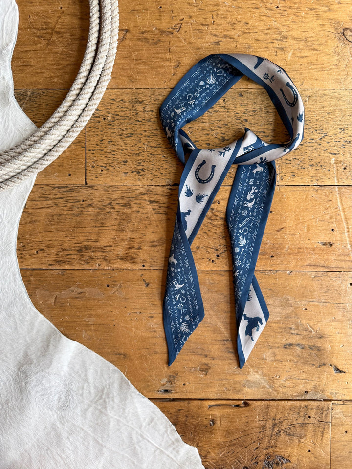 A Down South Navy Satin Twilly from Savannah Sevens western life{&}style, featuring a blue and white western print, lies on a wooden surface next to a coiled rope and white textured fabric.