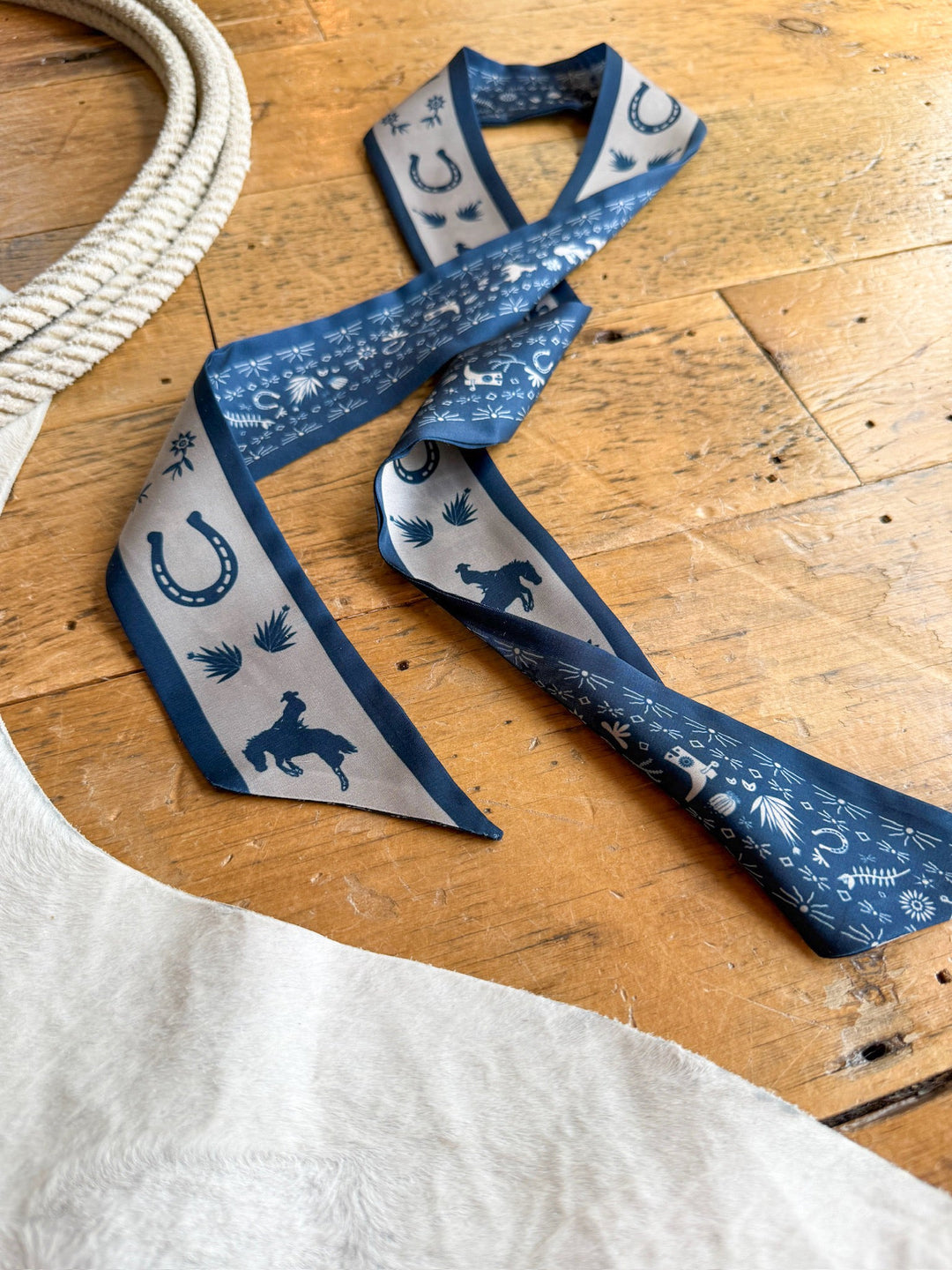 The Down South Navy Satin Twilly by Savannah Sevens western life{&}style, featuring a western print with horseshoes, cacti, and animal designs in shades of blue and beige, is displayed on a rustic wooden surface beside a rope and a light-colored cowhide.