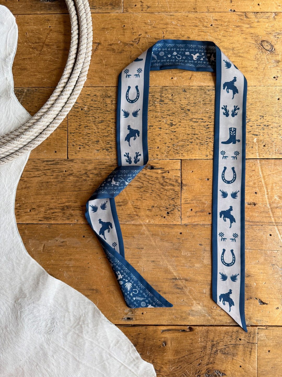 A Down South Navy Satin Twilly from Savannah Sevens western life{&}style, featuring cowboy-themed designs of horses, boots, hats, and horseshoes in blue and white, is displayed on a rustic wooden floor beside a coiled rope and a white animal hide.