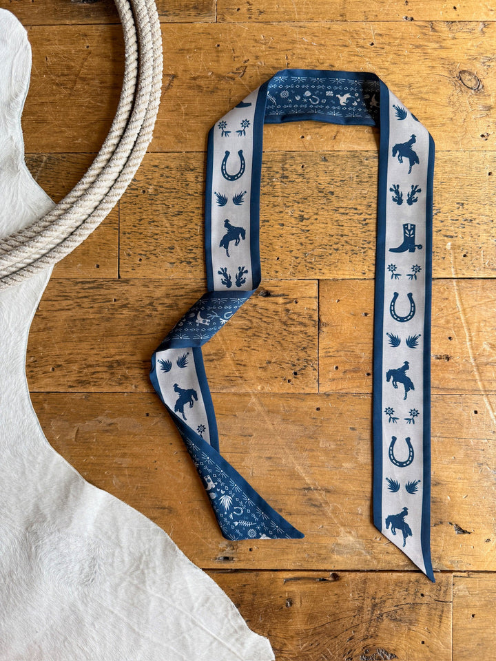 A Down South Navy Satin Twilly from Savannah Sevens western life{&}style, featuring cowboy-themed designs of horses, boots, hats, and horseshoes in blue and white, is displayed on a rustic wooden floor beside a coiled rope and a white animal hide.