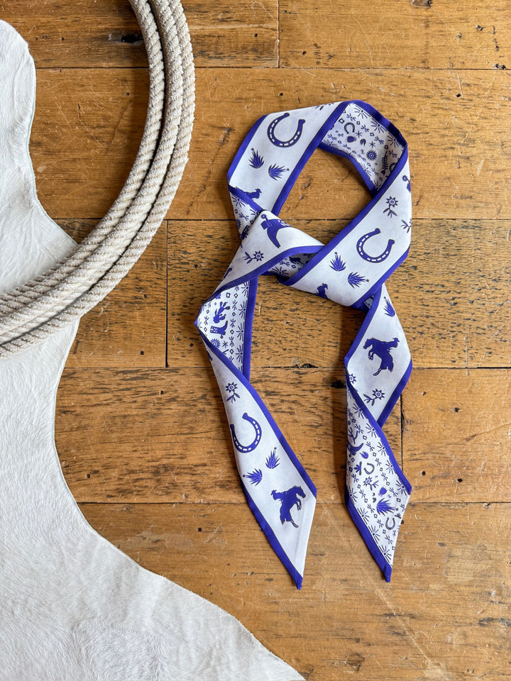 The Down South Royal Purple Satin Twilly by Savannah Sevens western life{&}style lies on a wooden floor, next to a coiled rope and the edge of a white cowhide rug.