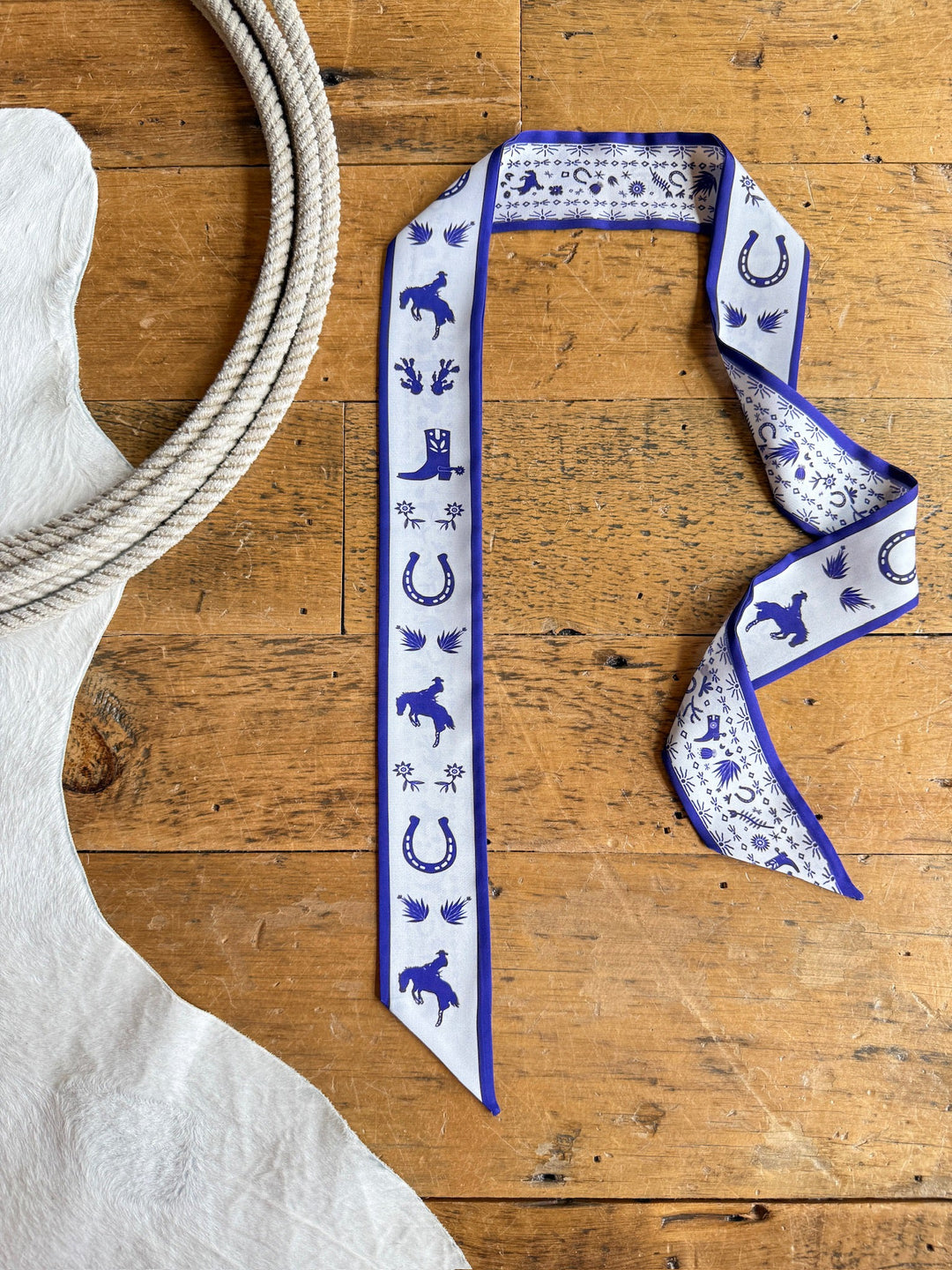 A Down South Royal Purple Satin Twilly from Savannah Sevens western life{&}style lies on a wooden floor next to a white cowhide rug and a coiled rope.