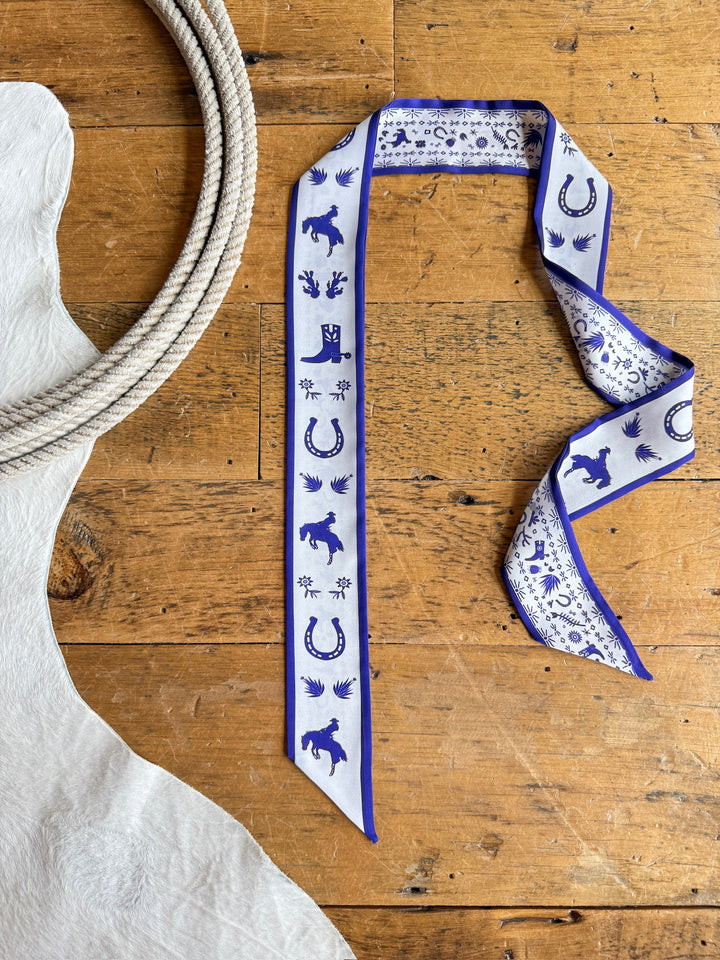 A Down South Royal Purple Satin Twilly from Savannah Sevens western life{&}style lies on a wooden floor next to a white cowhide rug and a coiled rope.