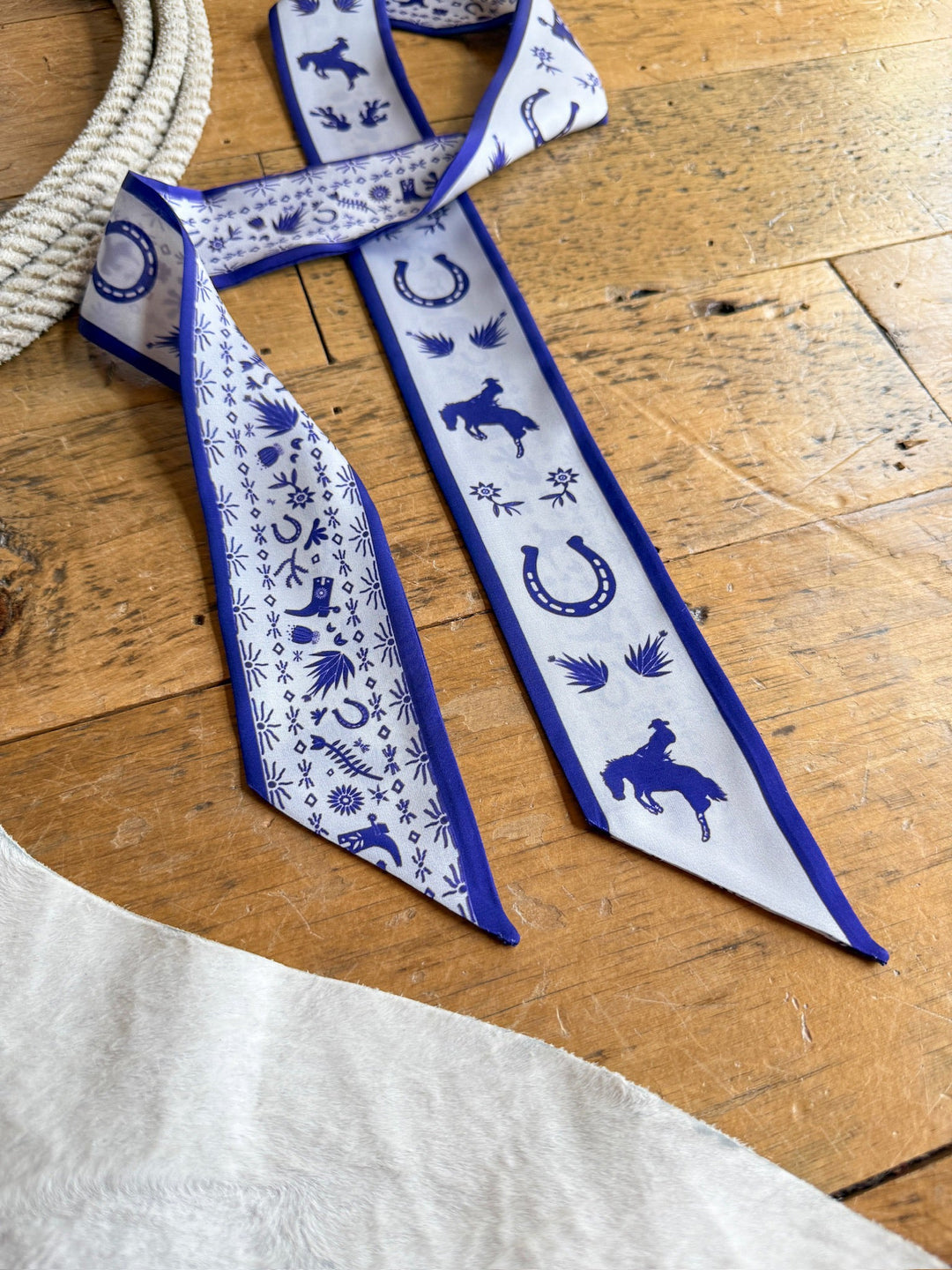 A Down South Royal Purple Satin Twilly by Savannah Sevens western life{&}style, featuring cowboy-themed designs such as horses, horseshoes, and cowboy hats, rests on a wooden surface beside a rope and a white textured fabric.