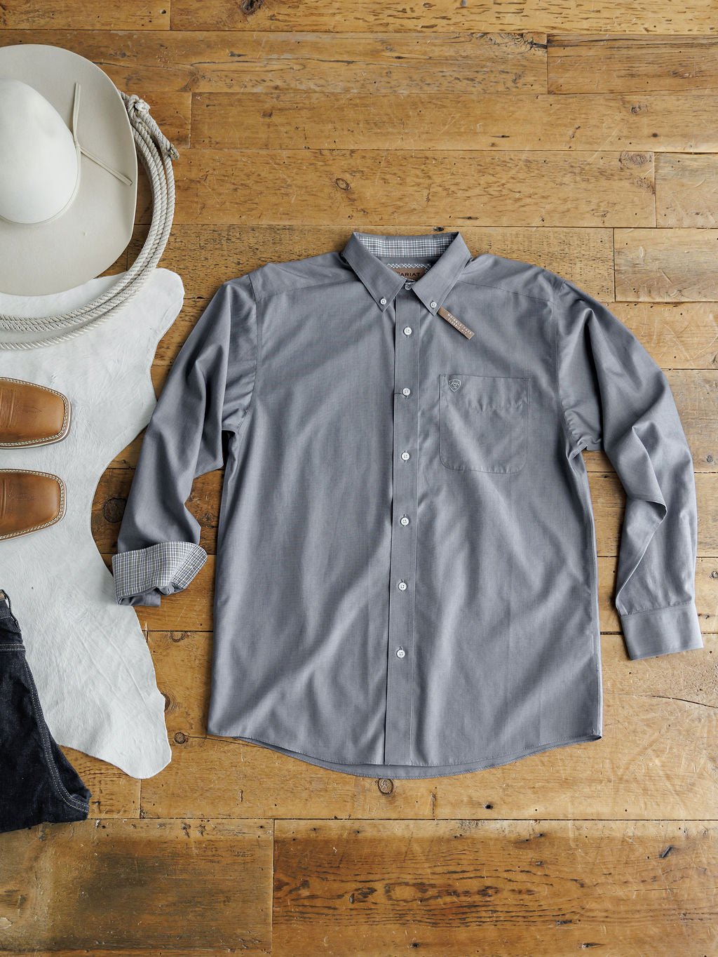 The Driggs Ariat Men's Slate Button Up from Savannah Sevens western life{&}style is laid flat on a wooden floor with a white cowboy hat, coiled rope, brown boots, and cowhide. This men's long sleeve shirt features wrinkle-free cotton for all-day style.