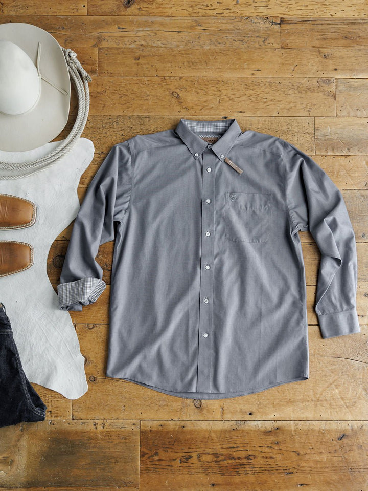 The Driggs Ariat Men's Slate Button Up from Savannah Sevens western life{&}style is laid flat on a wooden floor with a white cowboy hat, coiled rope, brown boots, and cowhide. This men's long sleeve shirt features wrinkle-free cotton for all-day style.