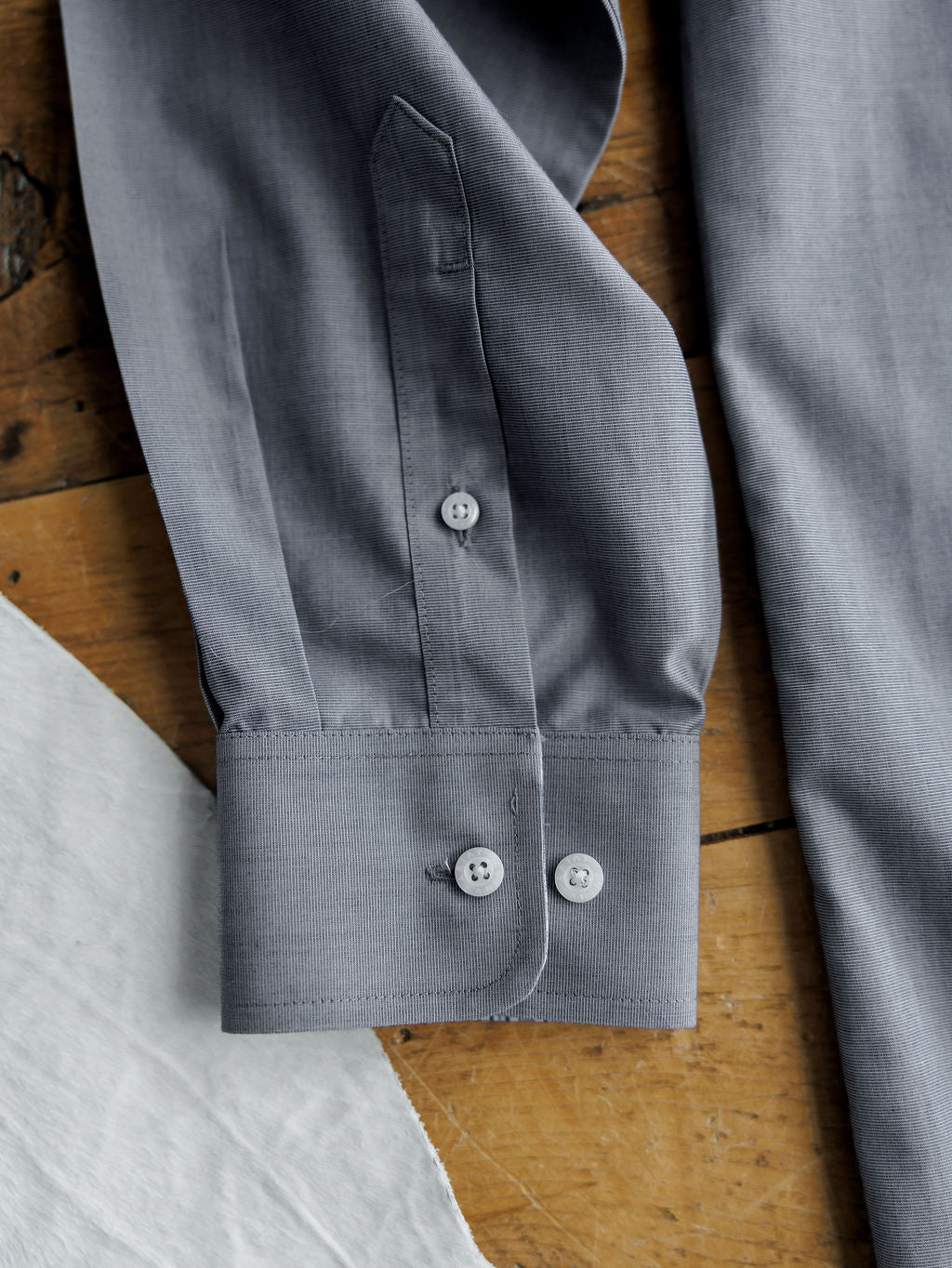 A close-up of the Driggs Ariat Men's Slate Button Up from Savannah Sevens western life{&}style features a gray cuff with two buttons on wood next to white fabric. Made from wrinkle-free cotton for easy care and a crisp look.