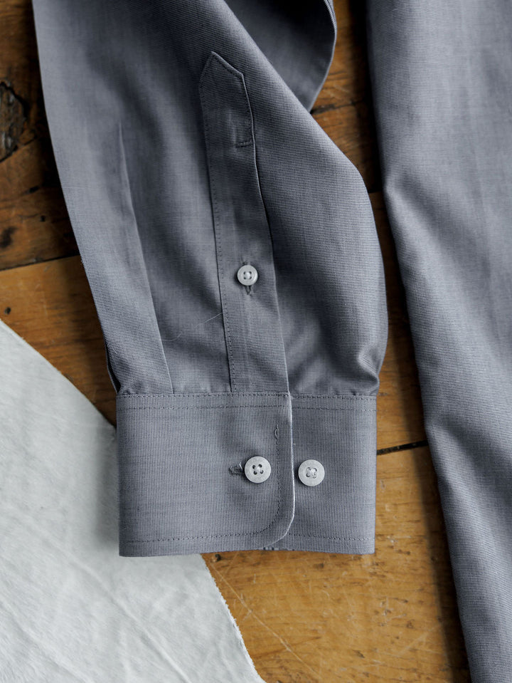A close-up of the Driggs Ariat Men's Slate Button Up from Savannah Sevens western life{&}style features a gray cuff with two buttons on wood next to white fabric. Made from wrinkle-free cotton for easy care and a crisp look.