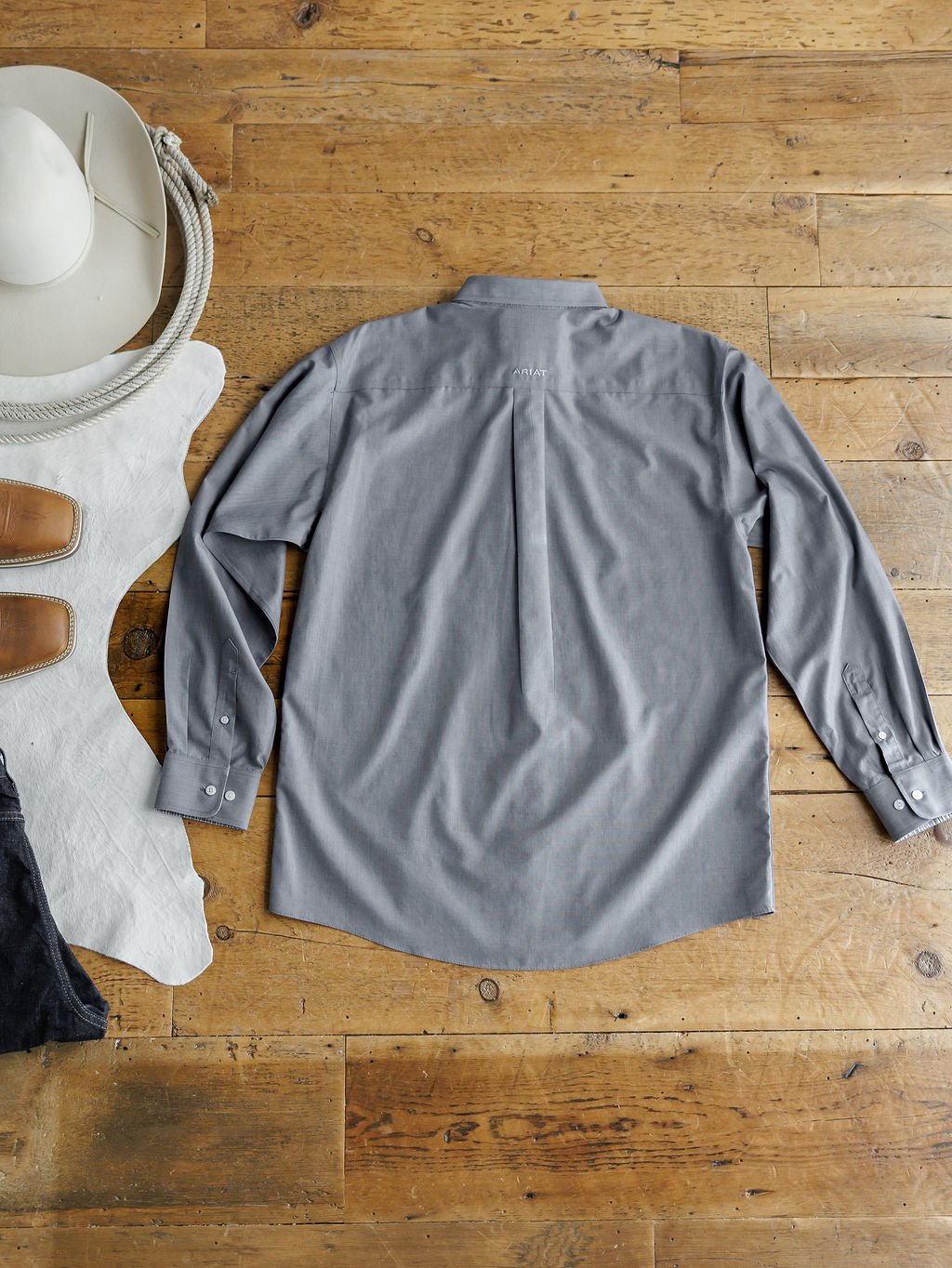 The Driggs Ariat Men's Slate Button Up by Savannah Sevens western life{&}style is laid flat, back up, on a wooden floor with a white cowboy hat, brown boots, and black pants beside it.