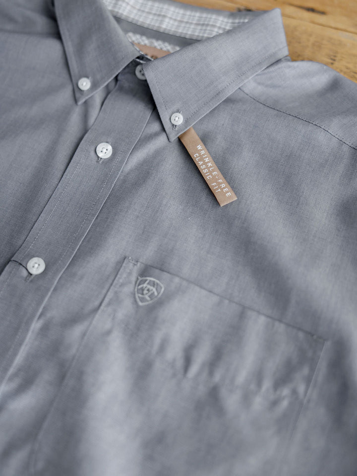A close-up of the Driggs Ariat Men's Slate Button Up from Savannah Sevens western life{&}style, neatly folded with a chest pocket, shield logo, and "Wrinkle Free Classic Fit" tag on the collar, is laid on a wooden surface.