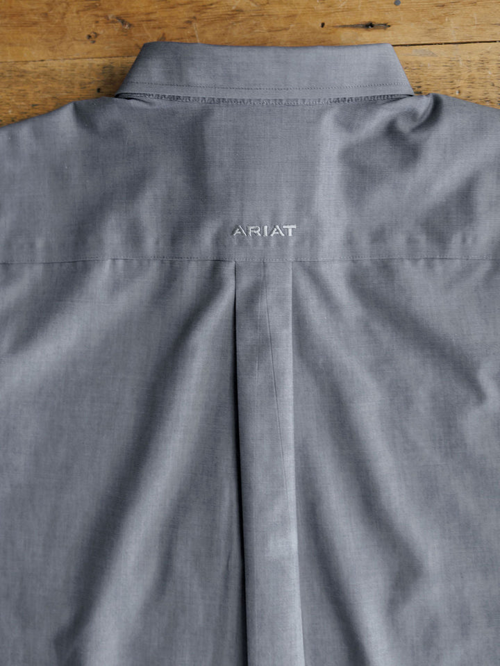The back view of the Driggs Ariat Men's Slate Button Up by Savannah Sevens western life{&}style shows the brand name ARIAT embroidered in white below the collar, displayed on a wooden surface.