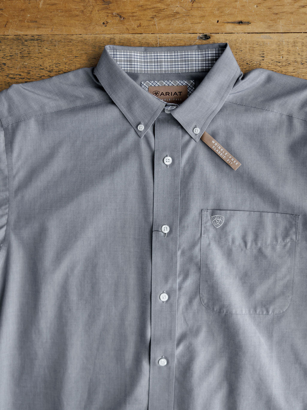 The Driggs Ariat Men's Slate Button Up by Savannah Sevens western life{&}style is a wrinkle-free cotton shirt with a chest pocket, button-down collar, white buttons, and a subtle logo on the pocket, laid neatly on a wooden surface.