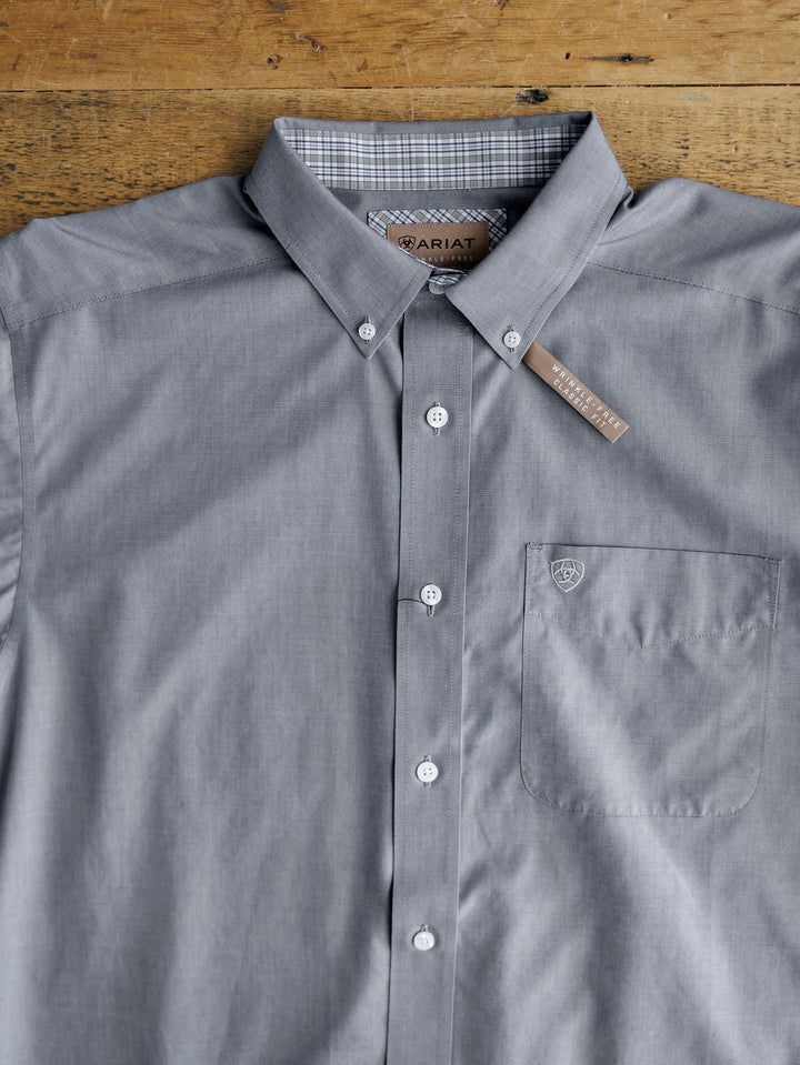The Driggs Ariat Men's Slate Button Up by Savannah Sevens western life{&}style is a wrinkle-free cotton shirt with a chest pocket, button-down collar, white buttons, and a subtle logo on the pocket, laid neatly on a wooden surface.
