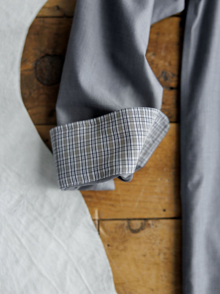 A close-up of the Driggs Ariat Men's Slate Button Up from Savannah Sevens western life{&}style shows a rolled cuff with a checkered pattern on wrinkle-free cotton, lying on wood near white fabric.