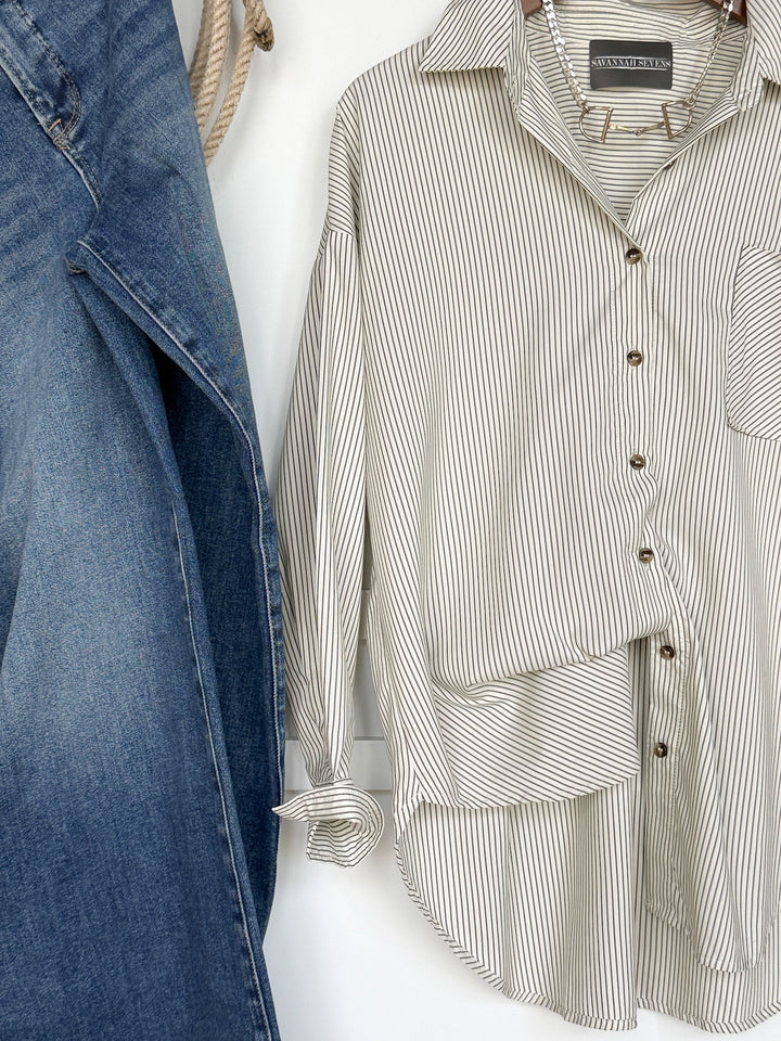 An oversized relaxed fit blue denim jacket hangs next to the Driscoll Pinstripe Button Up Top by Savannah Sevens western life{&}style, a white shirt featuring thin black pinstripes and a front pocket, both displayed on a white wall.