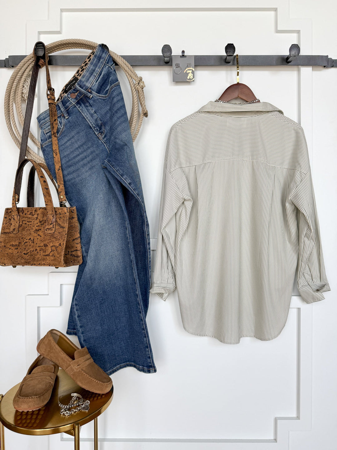 A Driscoll Pinstripe Button Up Top by Savannah Sevens western life{&}style, blue jeans, brown loafers, and a brown purse hang on hooks against a white wall; nearby, a rope and a brown belt are draped. The loafers rest on a small gold table.