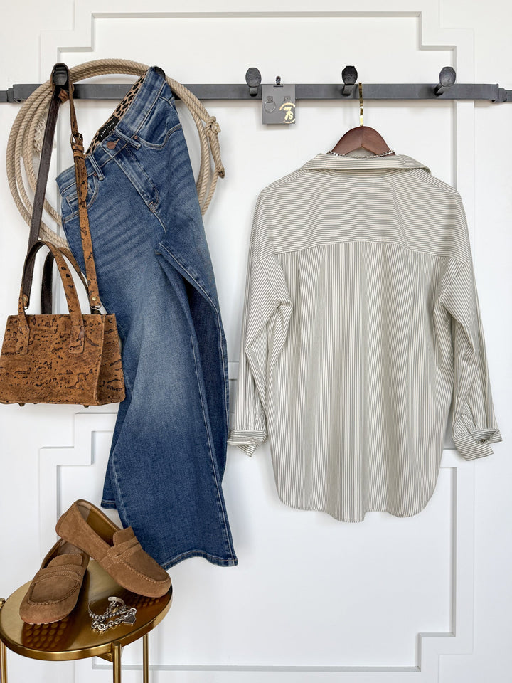 A Driscoll Pinstripe Button Up Top by Savannah Sevens western life{&}style, blue jeans, brown loafers, and a brown purse hang on hooks against a white wall; nearby, a rope and a brown belt are draped. The loafers rest on a small gold table.
