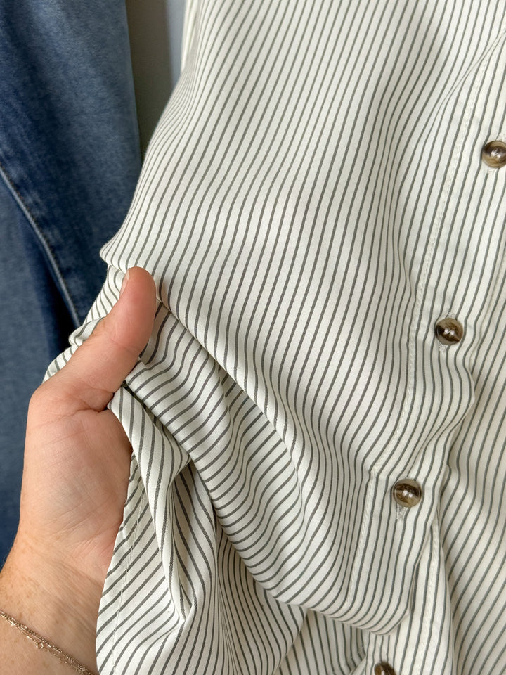 A hand holds the Driscoll Pinstripe Button Up Top by Savannah Sevens western life{&}style, a white shirt featuring thin black vertical pinstripes and metal buttons. A piece of denim fabric is visible in the background.