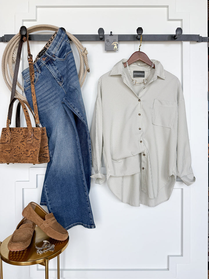 A Driscoll Pinstripe Button Up Top by Savannah Sevens western life{&}style, blue jeans, a brown textured handbag, tan moccasins, and a coiled rope hang neatly on a wall rack, with a small round table beneath the shoes.
