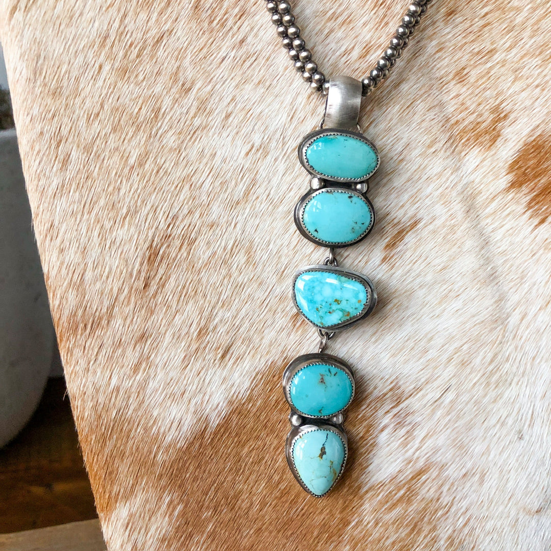 The Dry Creek Pendant by Savannah Sevens western life{&}style is a sterling silver necklace featuring four uniquely-shaped Dry Creek Turquoise stones in a vertical design, shown on a cowhide background.