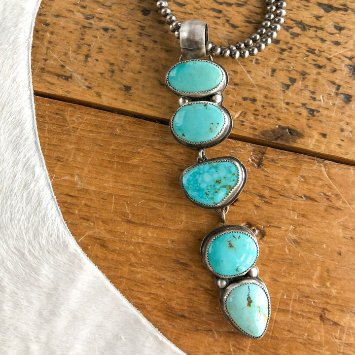 The Dry Creek Pendant by Savannah Sevens western life{&}style features five handmade turquoise stones set vertically in sterling silver bezels, shown on a wooden surface next to white fabric.