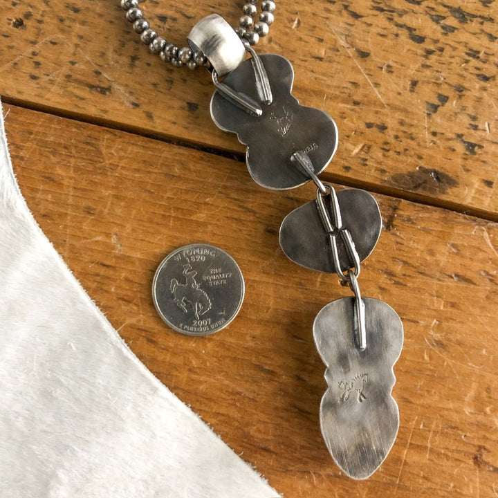 The Dry Creek Pendant by Savannah Sevens western life{&}style, a sterling silver necklace with three abstract links, is displayed on wood beside a U.S. quarter for scale and draped over white fabric in the corner.