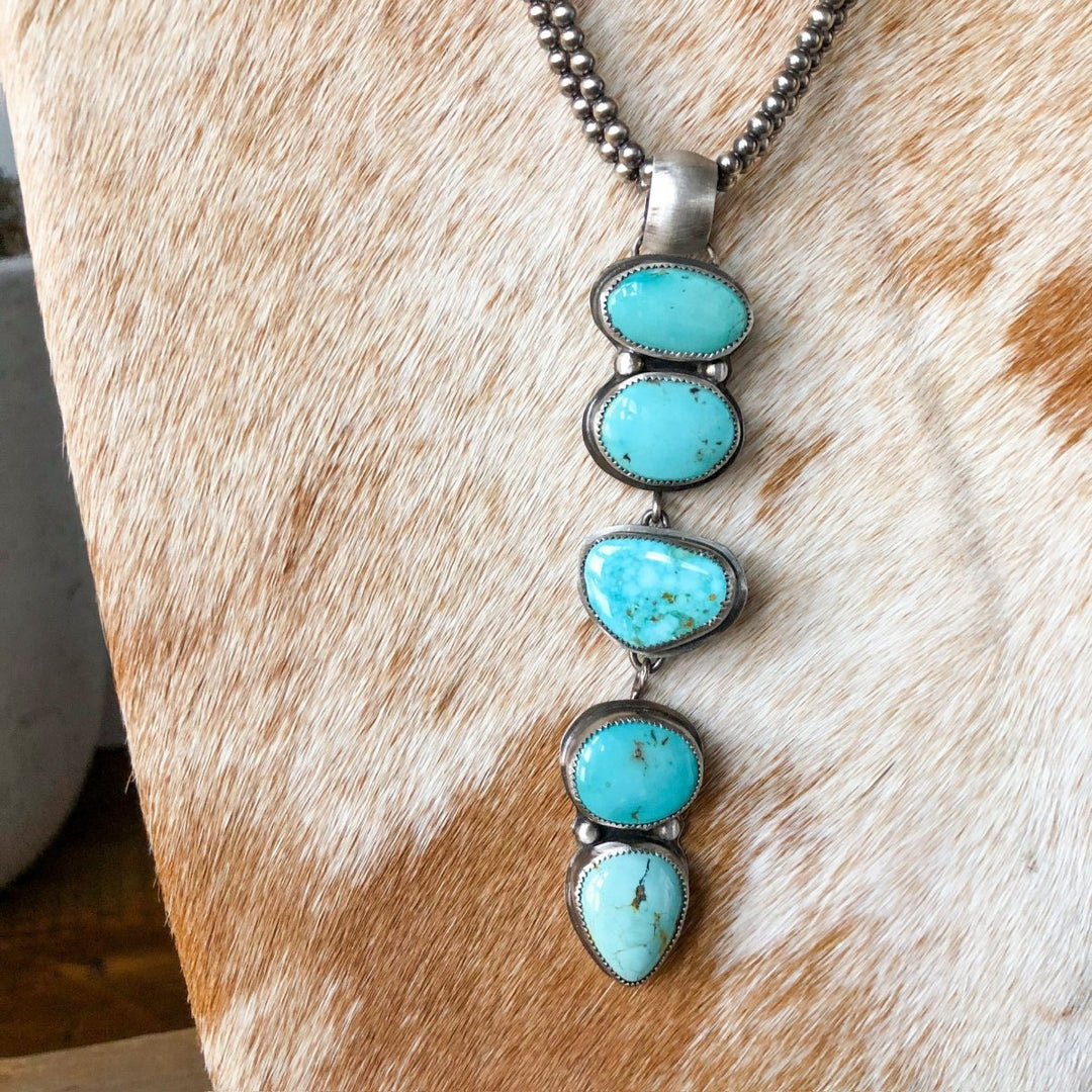 The Dry Creek Pendant by Savannah Sevens western life{&}style is a sterling silver necklace featuring four uniquely-shaped Dry Creek Turquoise stones in a vertical design, shown on a cowhide background.